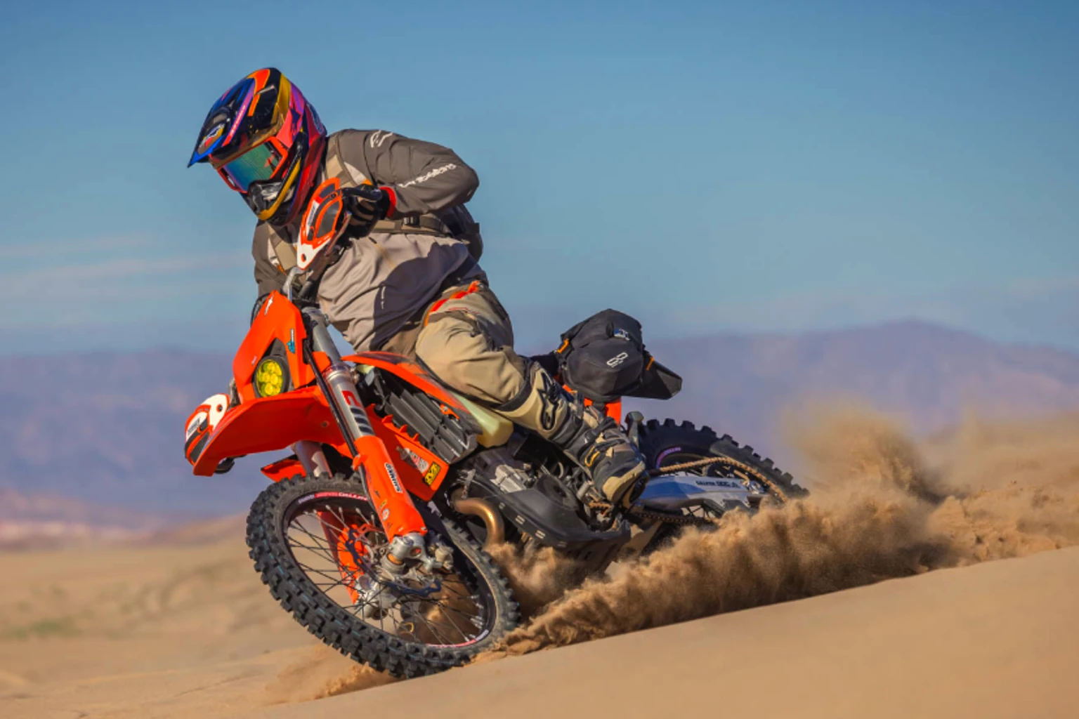 A fully equipped dual sport rider carving through a sandy desert turn with a wall of dust behind the motorcycle.